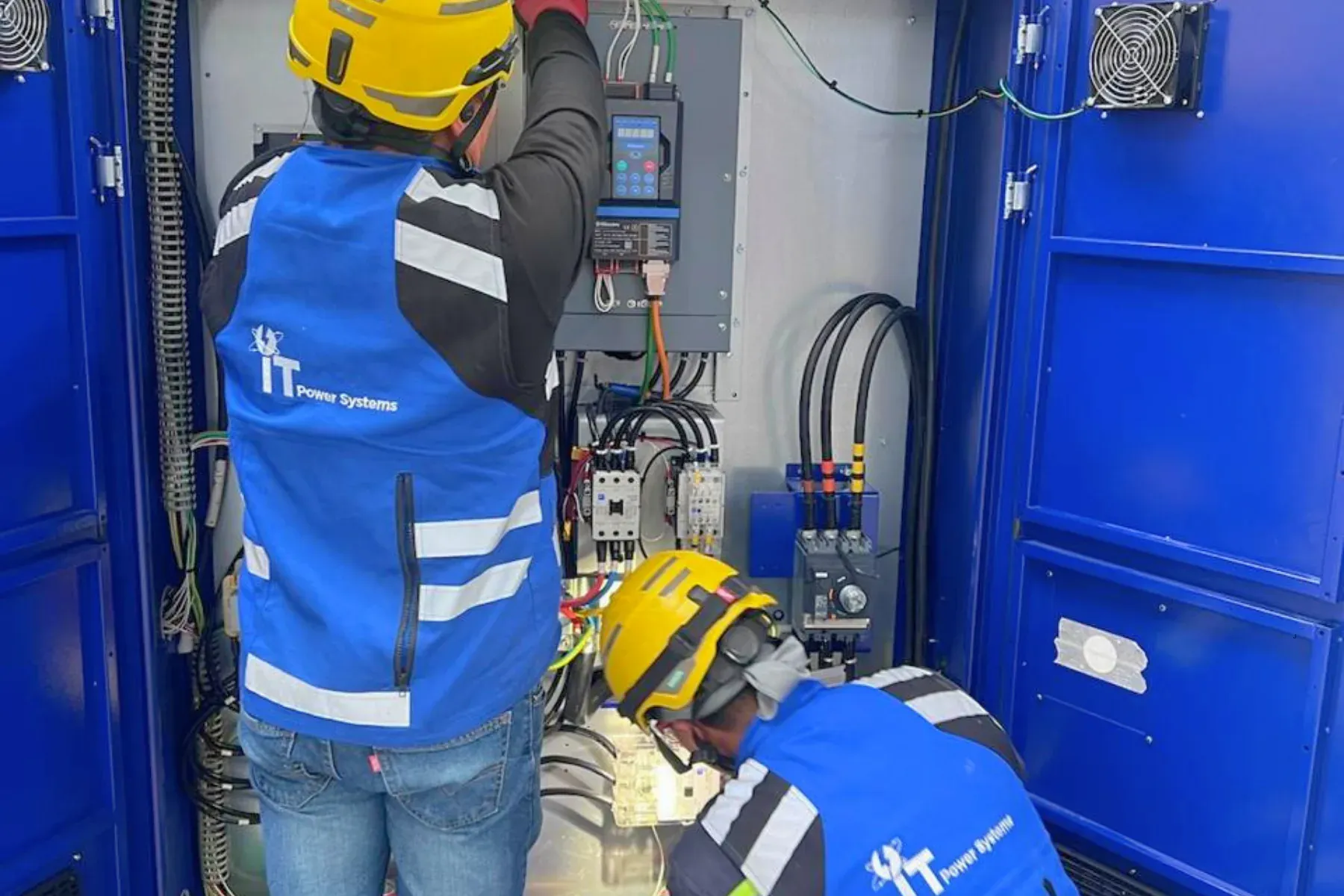 Dos técnicos de iT Power Systems con chaleco azul y casco cableando un variador de frecuencia y sus contactores dentro de un gabinete de fuerza para alimentar una máquina