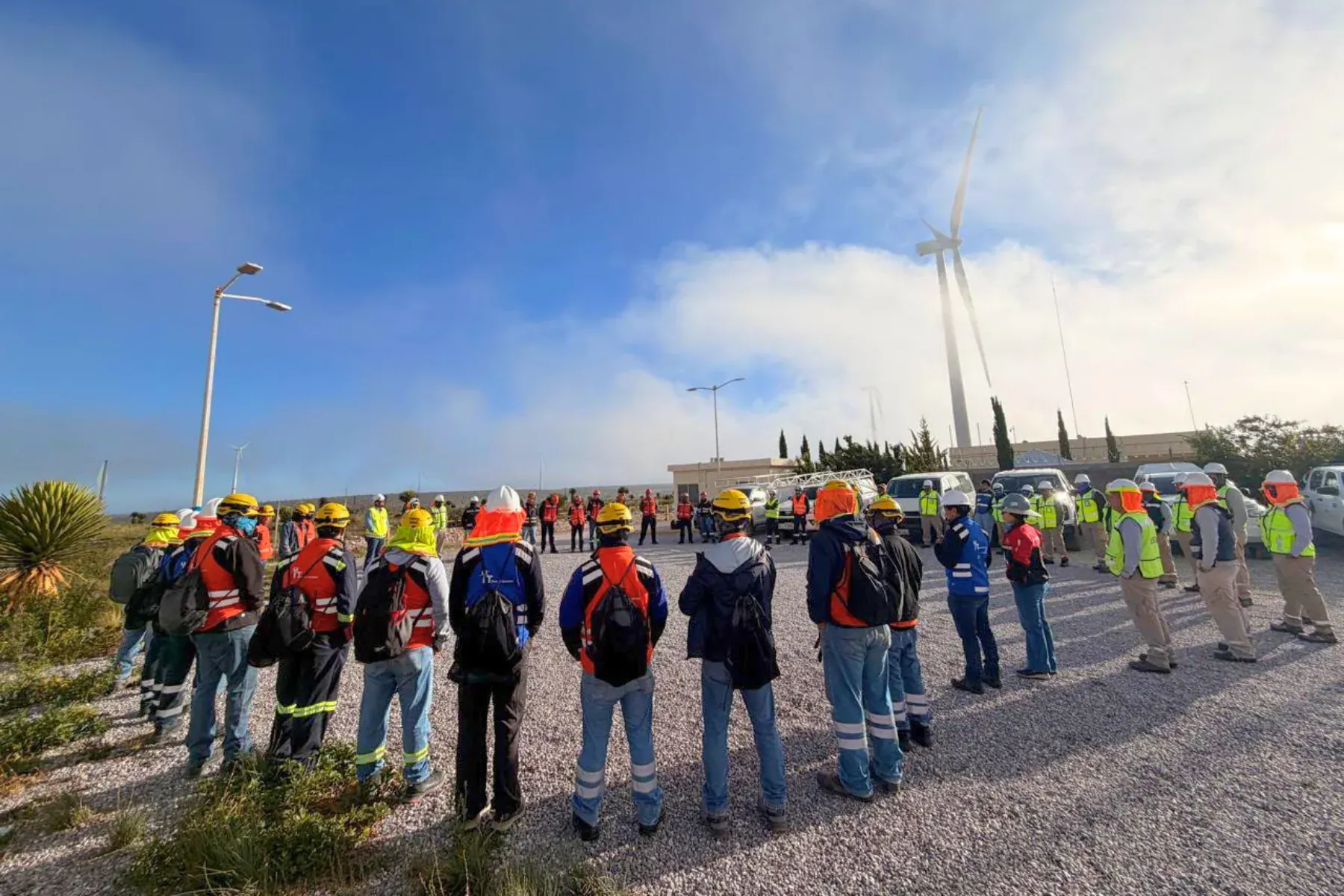 Cuadrilla de iT Power Systems en una charla de seguridad previa al trabajo, con casco y chaleco reflejante y un aerogenerador al fondo