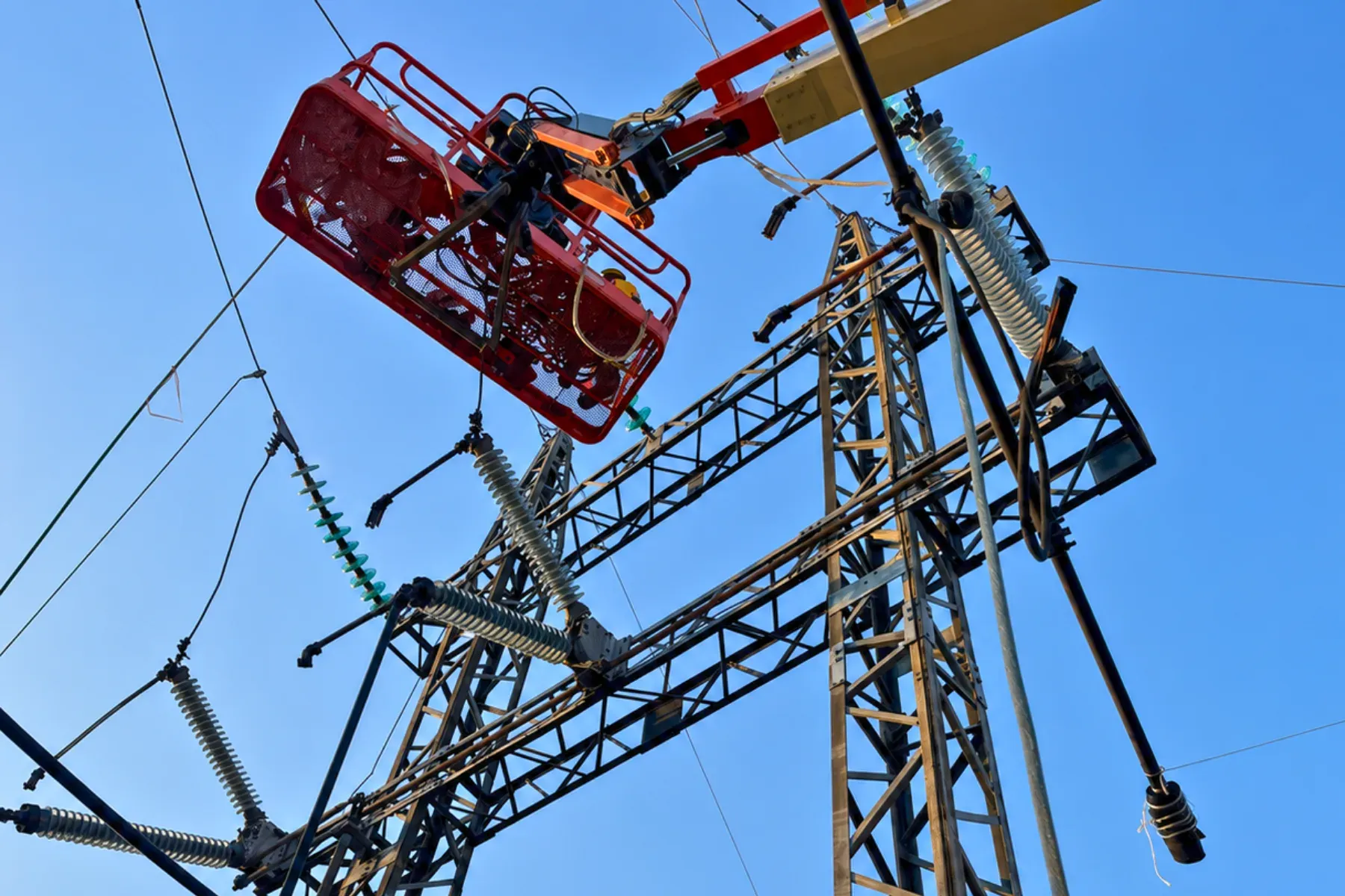 Canastilla elevadora con un trabajador junto a la estructura de una línea aérea de media tensión con aisladores y herrajes, durante la inspección y mantenimiento contra el cielo azul