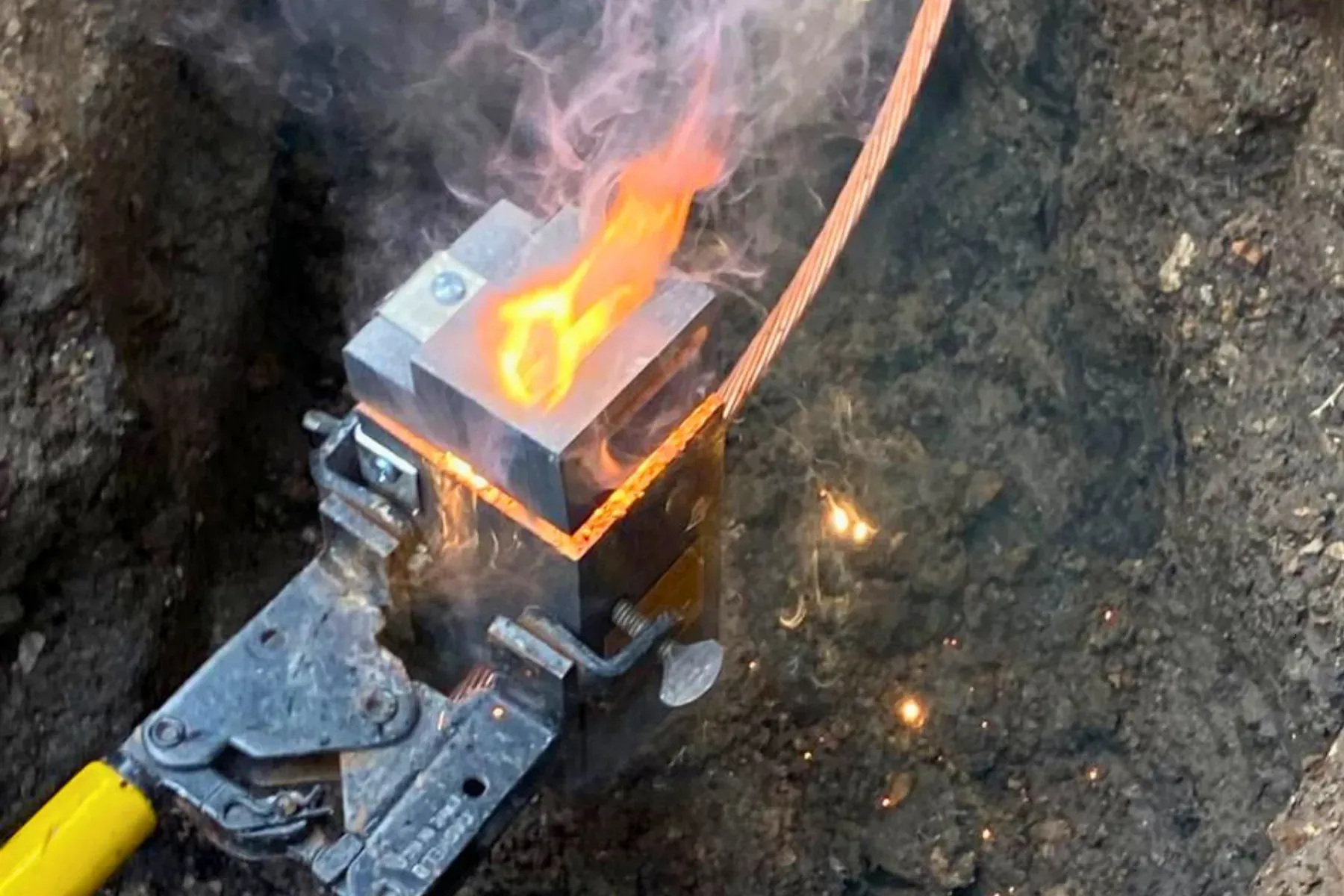 Soldadura exotérmica (cadweld) uniendo un cable de cobre desnudo de la malla de tierras dentro de una zanja, con el molde de grafito, la llama y las chispas de la reacción