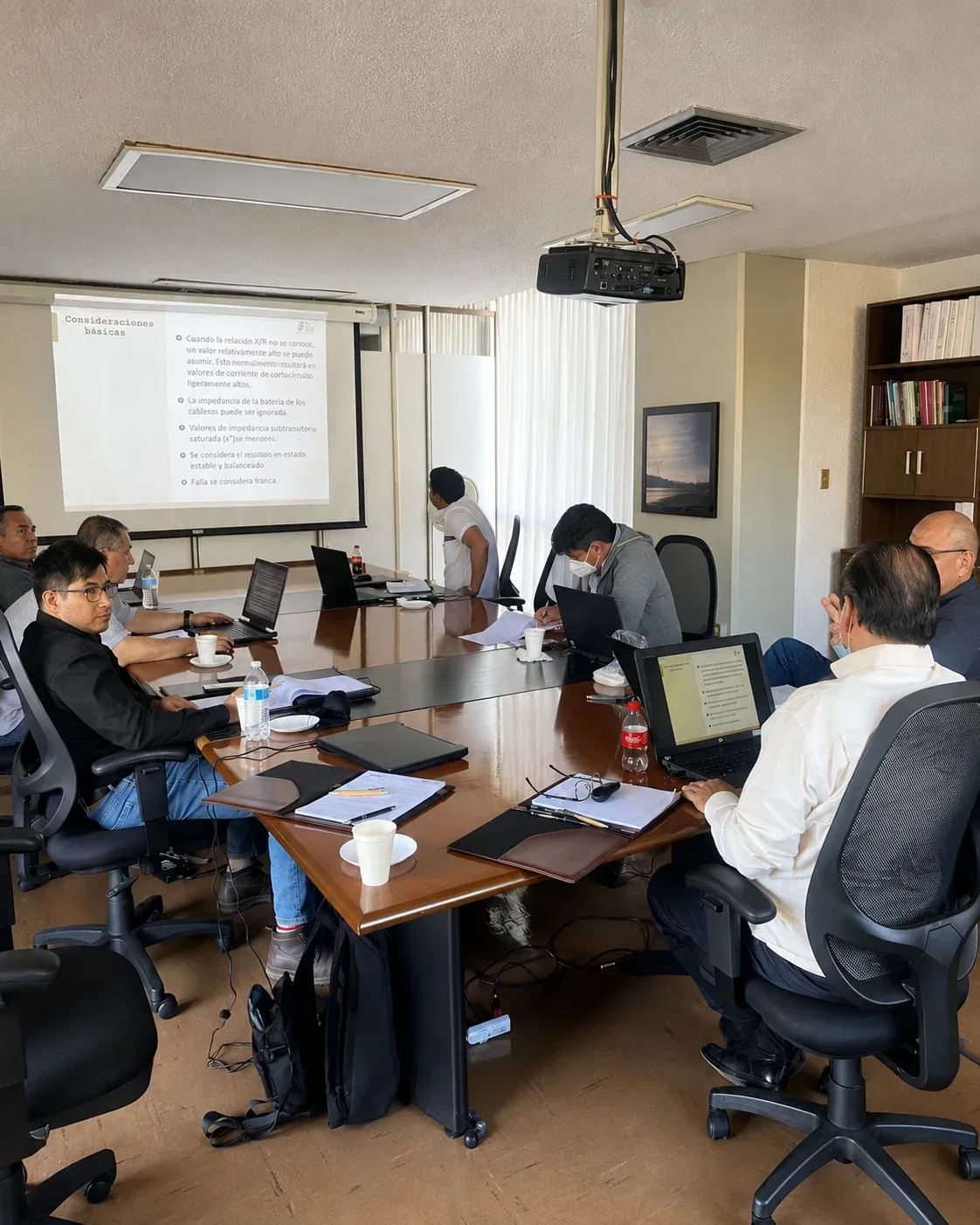 Reunión de ingeniería de iT Power Systems revisando en pantalla un estudio eléctrico con laptops alrededor de la mesa, en la fase de análisis previa a la ejecución
