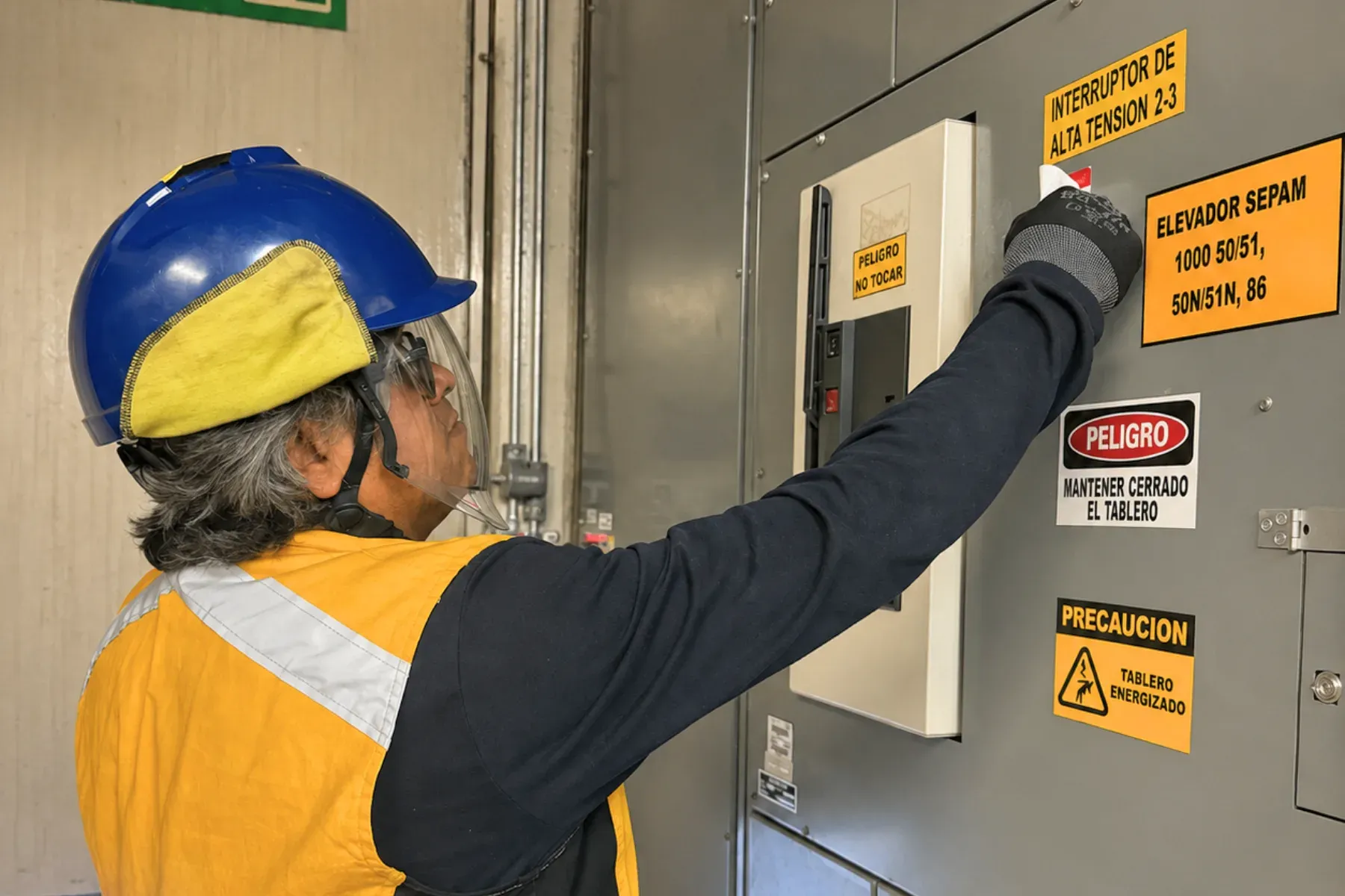 Técnico con casco, careta y guantes maniobrando el interruptor de un tablero de media tensión rotulado con etiquetas de seguridad de riesgo eléctrico y de tablero energizado