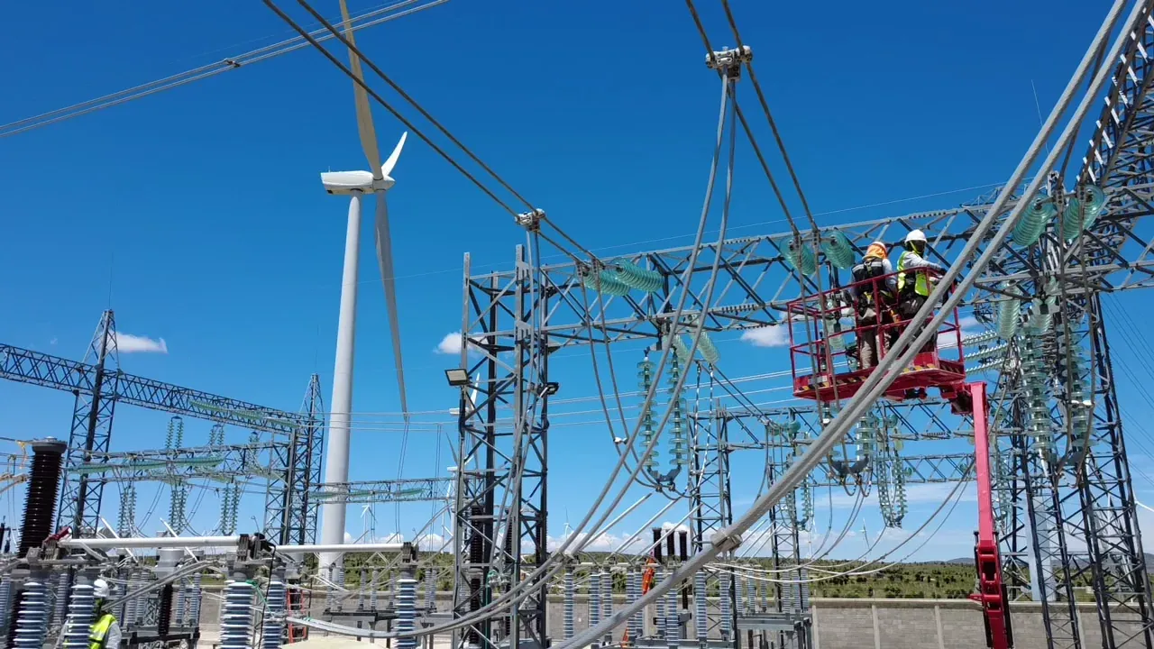 Cuadrilla de iT Power Systems trabajando en una subestación eléctrica de alta tensión, con operarios en canastilla y un aerogenerador al fondo