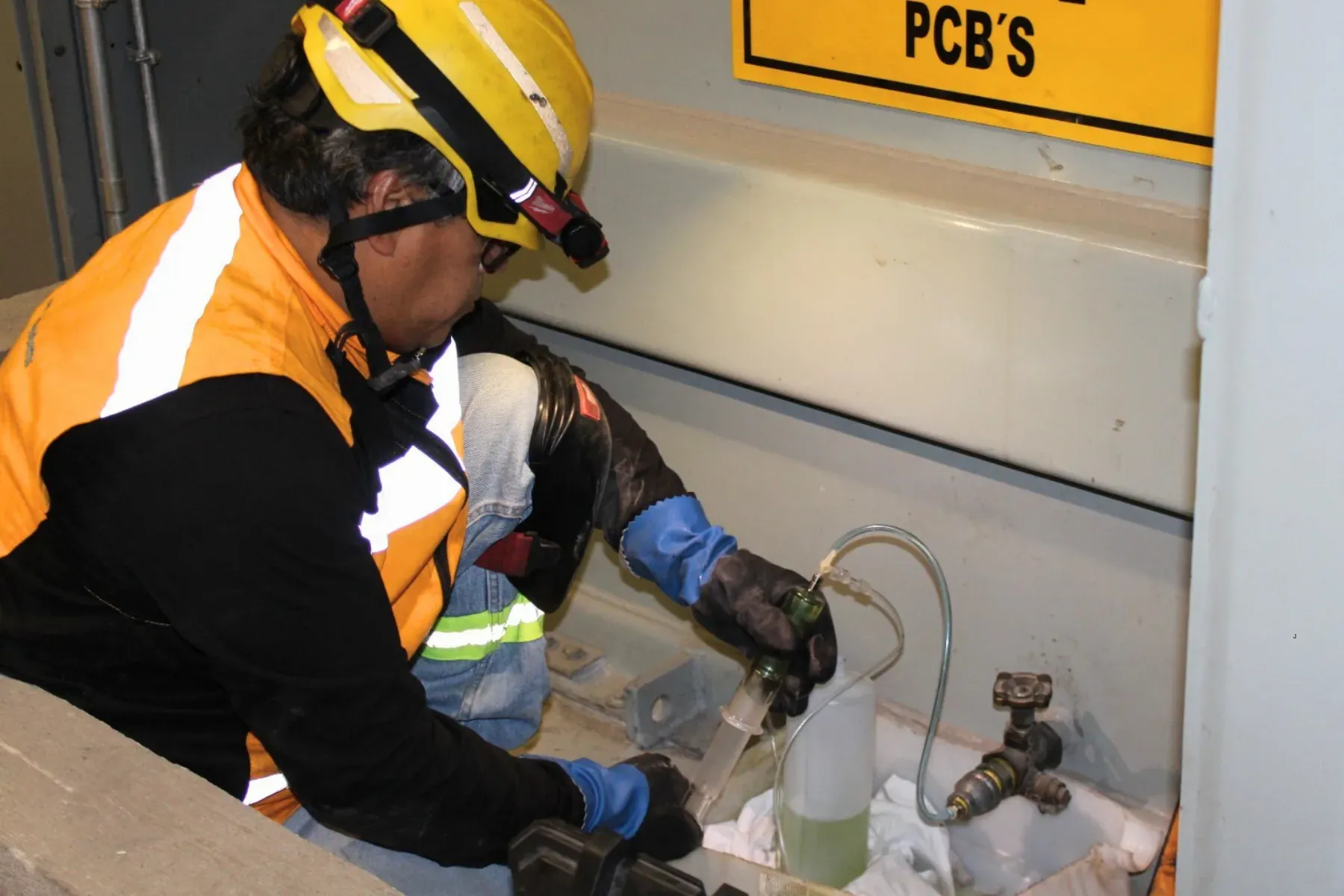 Técnico de iT Power Systems tomando una muestra de aceite aislante desde la válvula de un transformador con una jeringa de vidrio, junto a una placa de LIBRE DE BPC