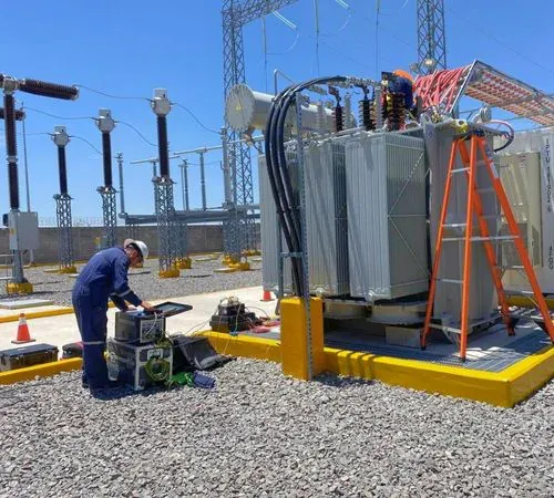 Dos técnicos de iT Power Systems trabajando en una subestación: uno con equipo de medición a pie de transformador y otro sobre una escalera apoyada en el tanque