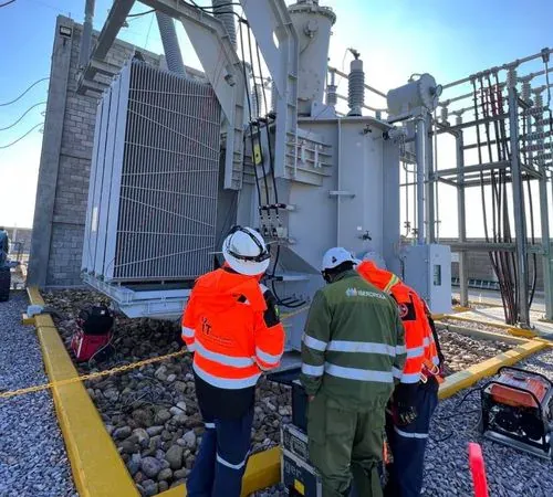 Cuadrilla de iT Power Systems y personal de planta frente a un transformador de potencia con radiadores, con equipo de prueba y planta de energía en el suelo