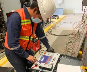 Ingeniero de iT Power Systems con chaleco reflejante ajustando un equipo de prueba portátil conectado a un juego de cables de potencia dentro de una nave industrial