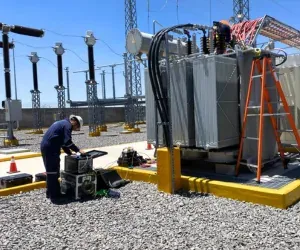 Ingeniero de iT Power Systems operando un equipo de prueba en el suelo junto a un transformador de potencia sobre su base, con la estructura de alta tensión de la subestación al fondo