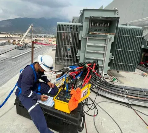 Ingeniero de iT Power Systems registrando lecturas junto a un equipo de prueba conectado con cables de alta corriente a un transformador seco, con la sierra al fondo
