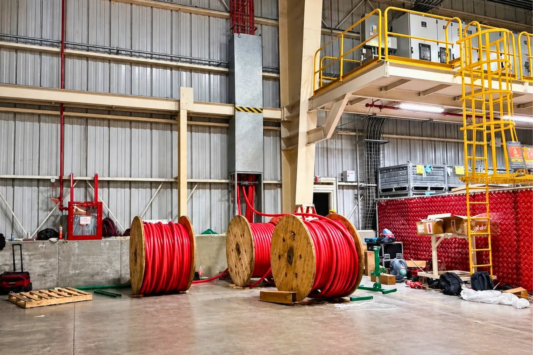 Carretes de madera con cable de potencia de media tensión en el piso de una nave industrial durante el montaje de una subestación, con el tablero instalado en una plataforma elevada al fondo